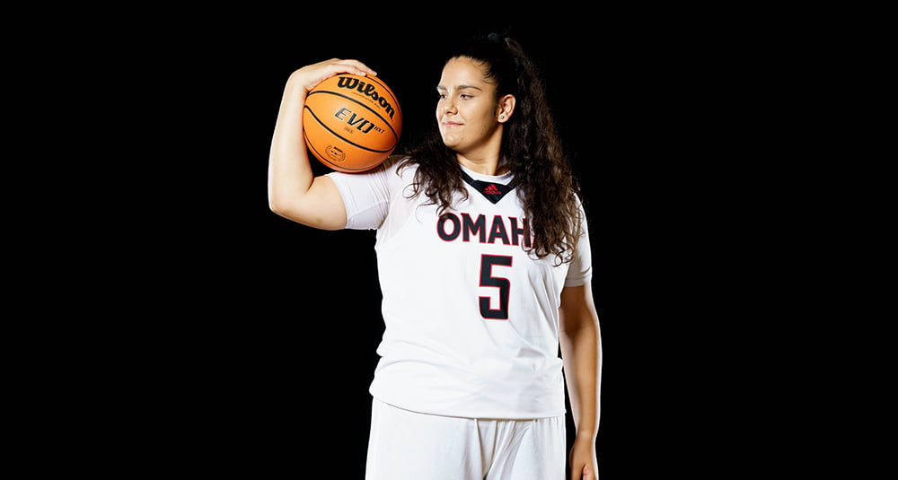 UNO graduate Elena Pilakouta holds a basketball in her flexed arm while wearing her white uniform